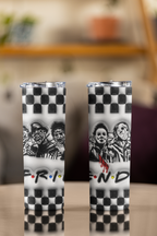 Two tall skinny tumblers with clear lids, featuring a black and white checkerboard pattern, horror character illustrations, and a stylized 'FRIENDS' logo, displayed on a table with a blurred indoor background.