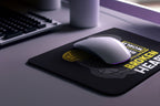 White mouse on a black mouse pad with text, placed on a desk with a keyboard and mug.
