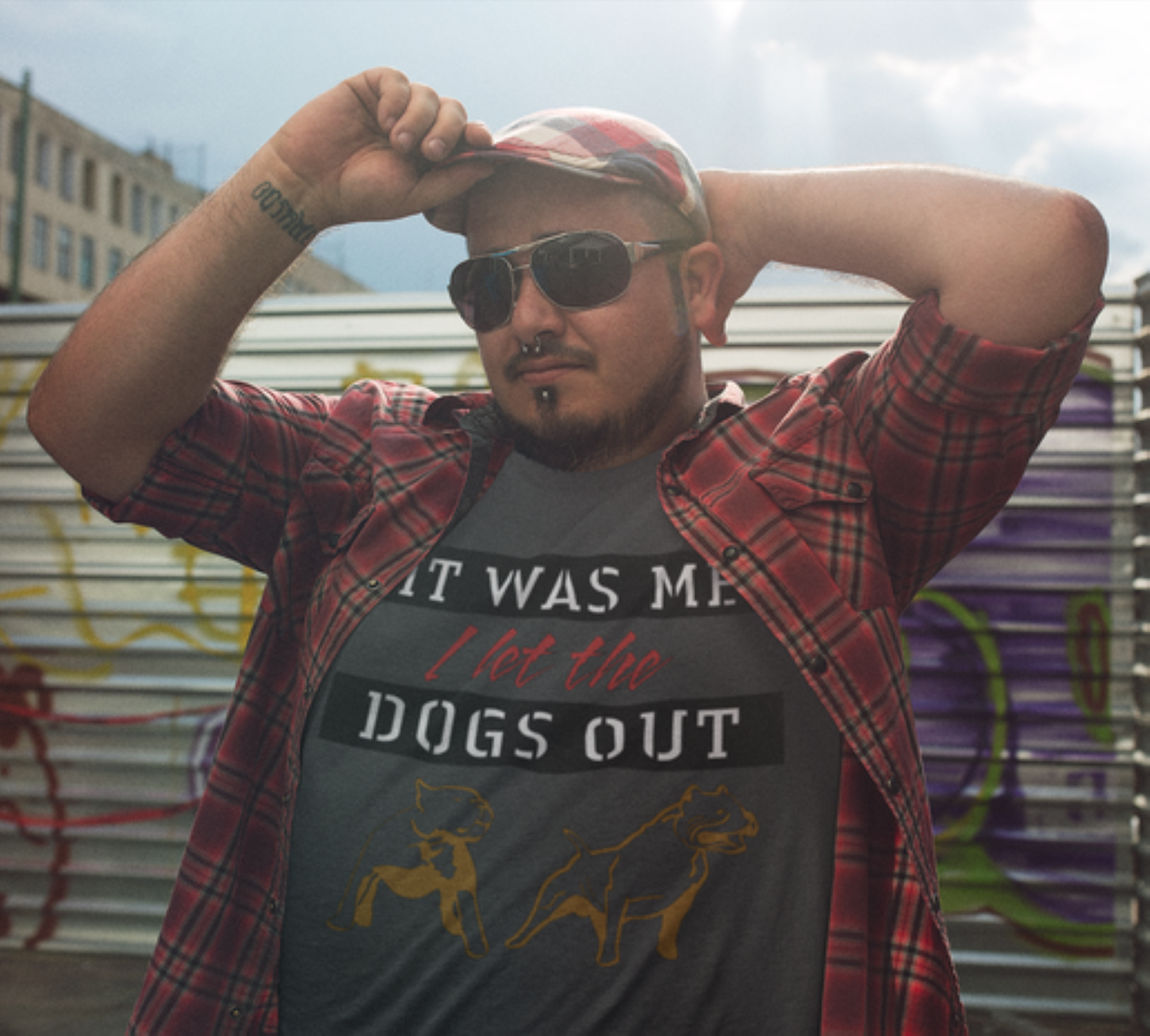 Man wearing a plaid shirt over a graphic t-shirt with text and dog illustrations, standing against a graffiti-covered wall.