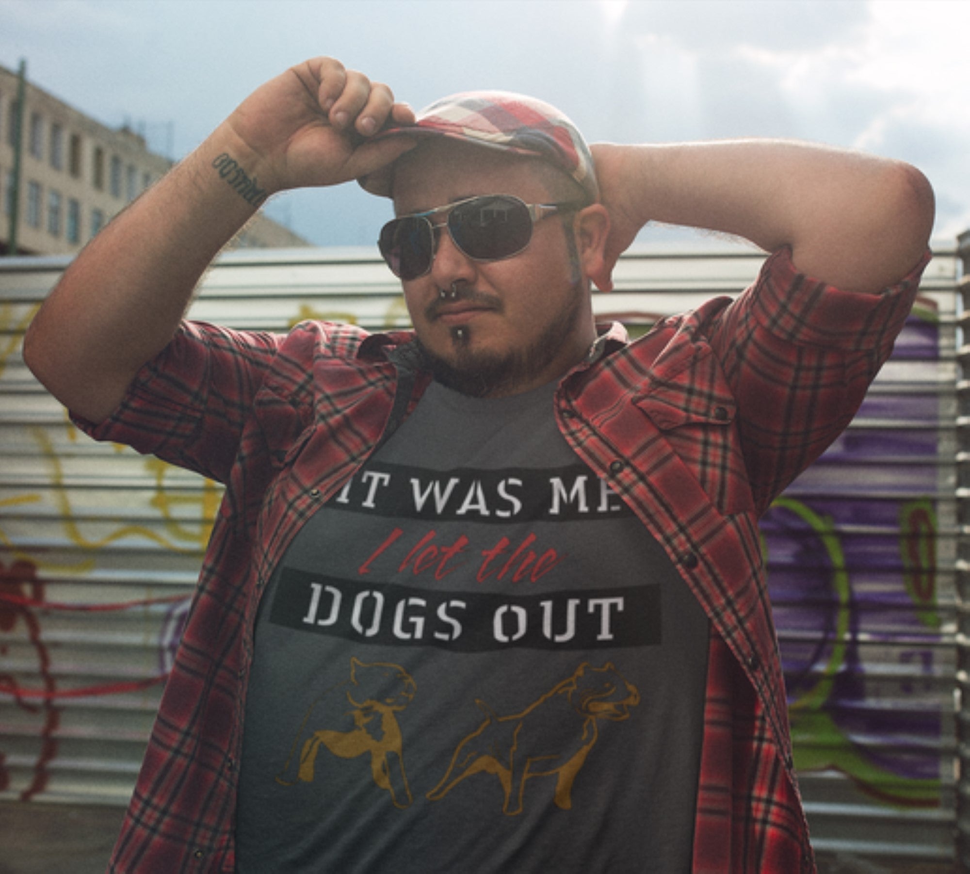 Man wearing a plaid shirt over a graphic t-shirt with text and dog illustrations, standing against a graffiti-covered wall.