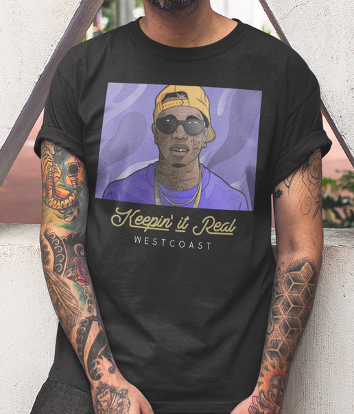 A man wearing a black graphic t-shirt with a purple and yellow print of a person in a cap and sunglasses, with the text 'Keepin' it Real WESTCOAST' below. The man has heavily tattooed arms and is leaning against a light-colored wall.