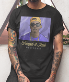 A man wearing a black graphic t-shirt with a purple and yellow print of a person in a cap and sunglasses, with the text 'Keepin' it Real WESTCOAST' below. The man has heavily tattooed arms and is leaning against a light-colored wall.