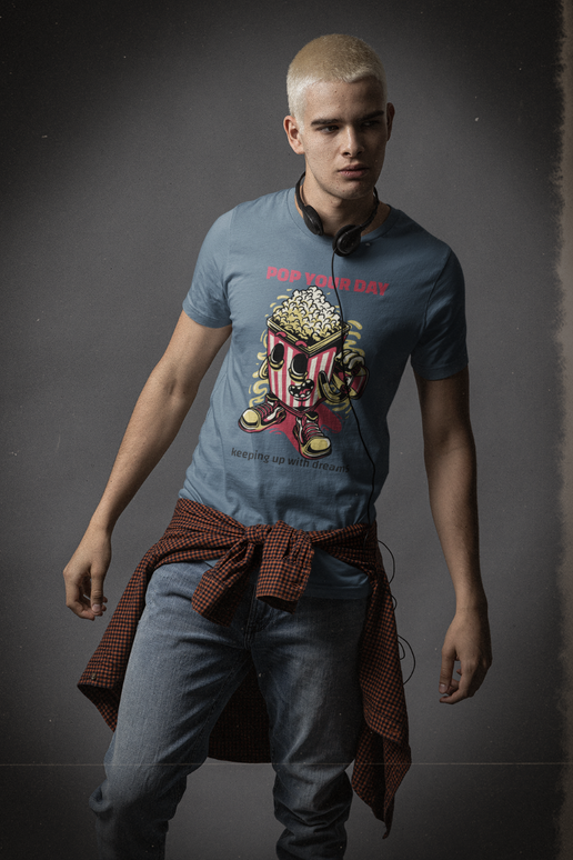 A man wearing a blue graphic t-shirt with a red and yellow popcorn character and the text 'POP YOUR DAY', paired with blue jeans and a brown shirt tied around his waist, set against a dark gray studio background.