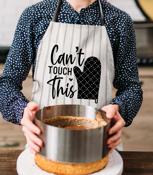 Person wearing an apron with 'Can't Touch This' text and an oven mitt graphic, holding a metal container.