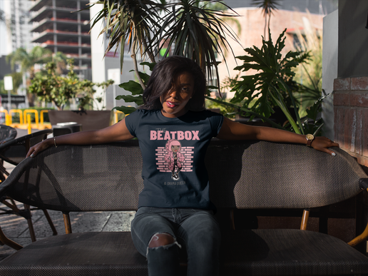 Woman wearing a 'Beatbox' t-shirt sitting on a bench outdoors with plants and buildings in the background.