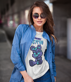 A woman wearing a graphic t-shirt with a purple bear and the text 'IT'S NOT BOY, IT'S Psycho', layered under a blue denim shirt and paired with sunglasses, set against a blurred urban background.