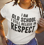 A person wearing a white graphic T-shirt with bold black text reading 'I AM OLD SCHOOL I STILL BELIEVE IN RESPECT', paired with blue jeans in a warm outdoor setting.