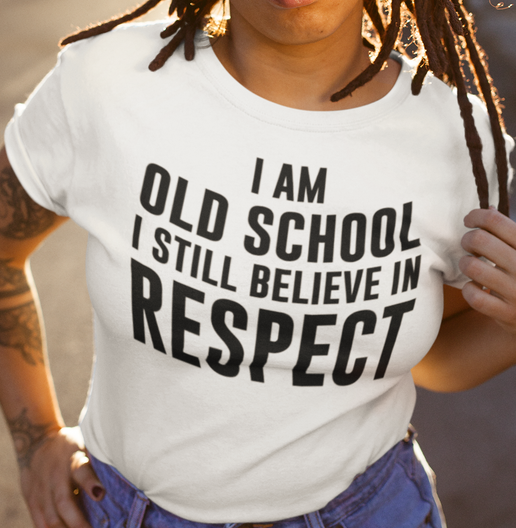 A person wearing a white graphic T-shirt with bold black text reading 'I AM OLD SCHOOL I STILL BELIEVE IN RESPECT', paired with blue jeans in a warm outdoor setting.