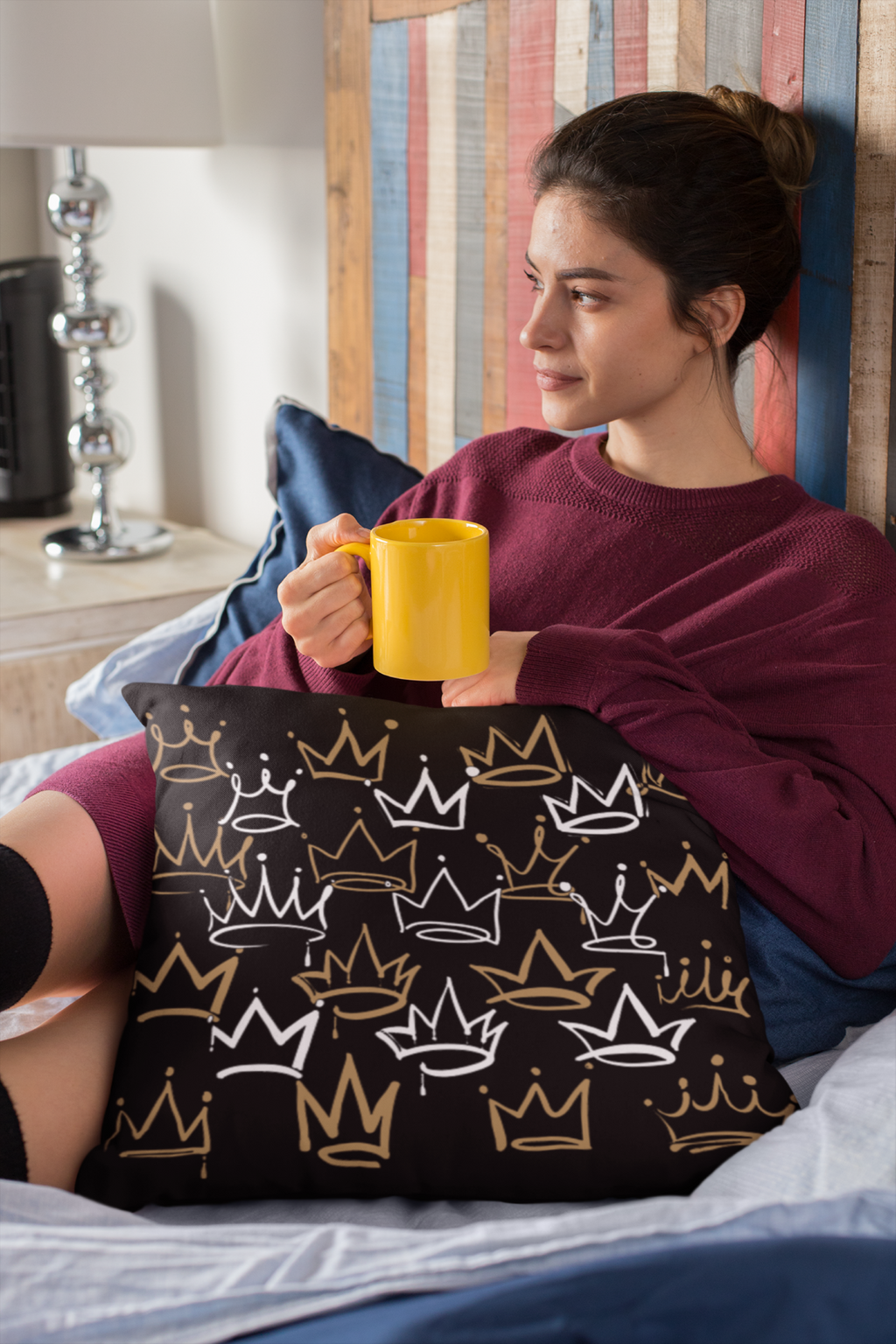 Woman holding a yellow mug on a pillow with crown patterns in a bedroom setting.