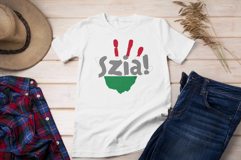 White t-shirt with 'Szia!' text and green design on a wooden surface with jeans, plaid shirt, and hat.