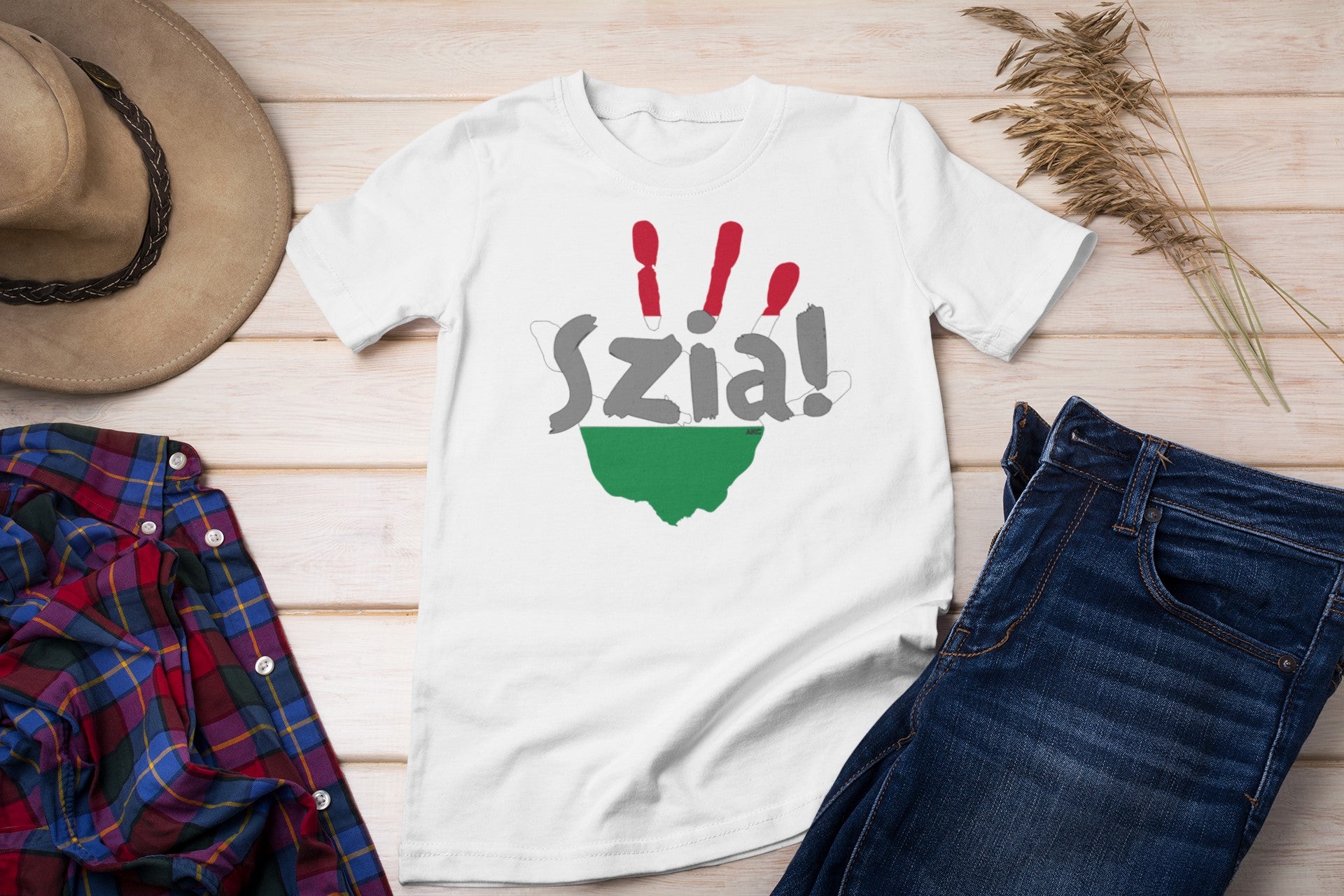 White t-shirt with 'Szia!' text and green design on a wooden surface with jeans, plaid shirt, and hat.
