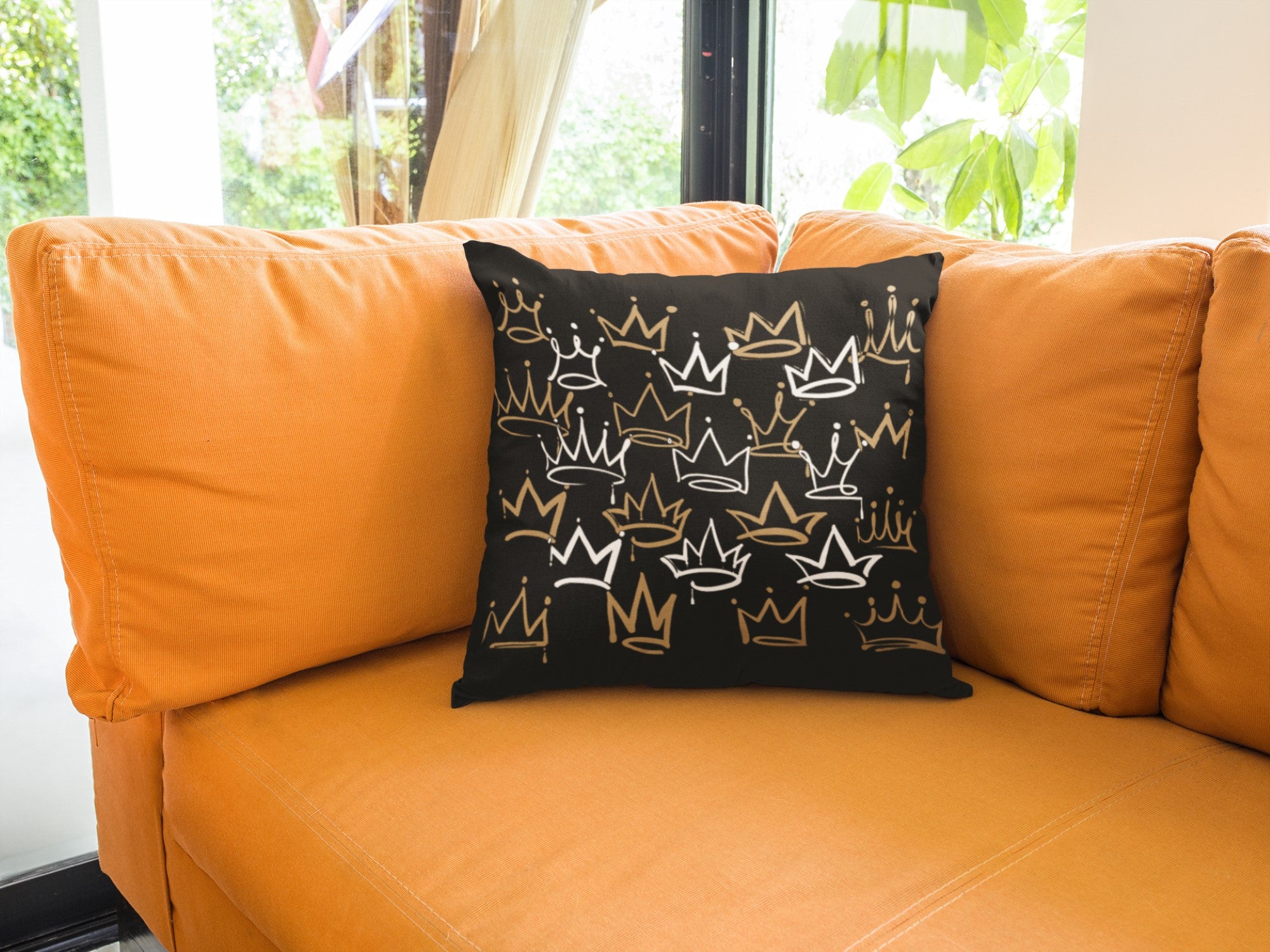Black pillow with gold crowns on an orange couch by a window