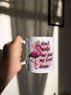 A hand holds a white ceramic mug with a pink flamingo graphic and the text 'don't make me put my foot down' against a softly lit wall with natural shadows.