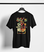 A black short-sleeve t-shirt hanging on a black hanger against a light gray wall. The shirt features a colorful graphic print on the front that reads '80's BABY 90's MADE ME' in a retro patterned font above an illustration of a cassette tape.