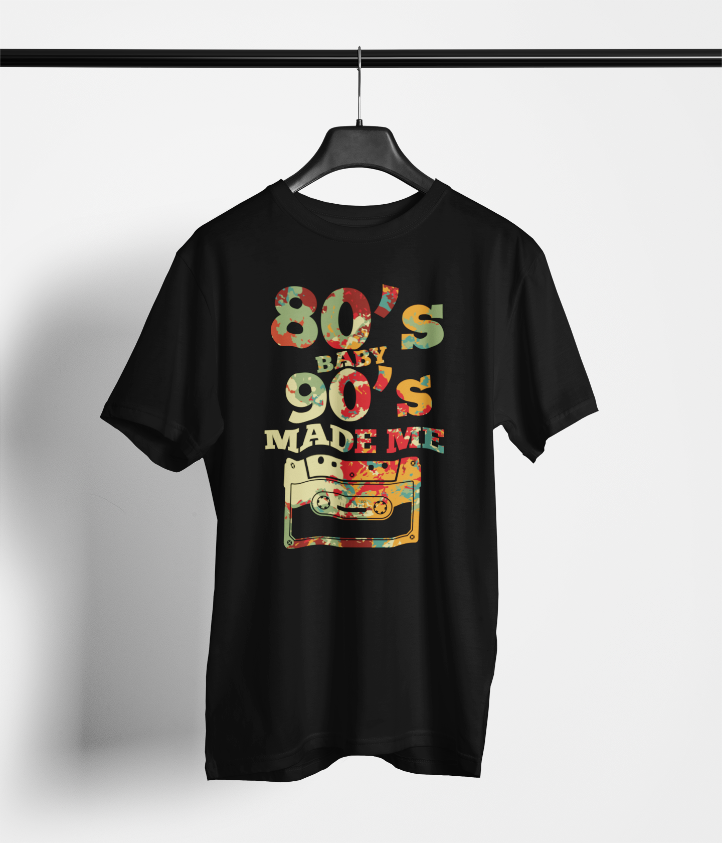 A black short-sleeve t-shirt hanging on a black hanger against a light gray wall. The shirt features a colorful graphic print on the front that reads '80's BABY 90's MADE ME' in a retro patterned font above an illustration of a cassette tape.