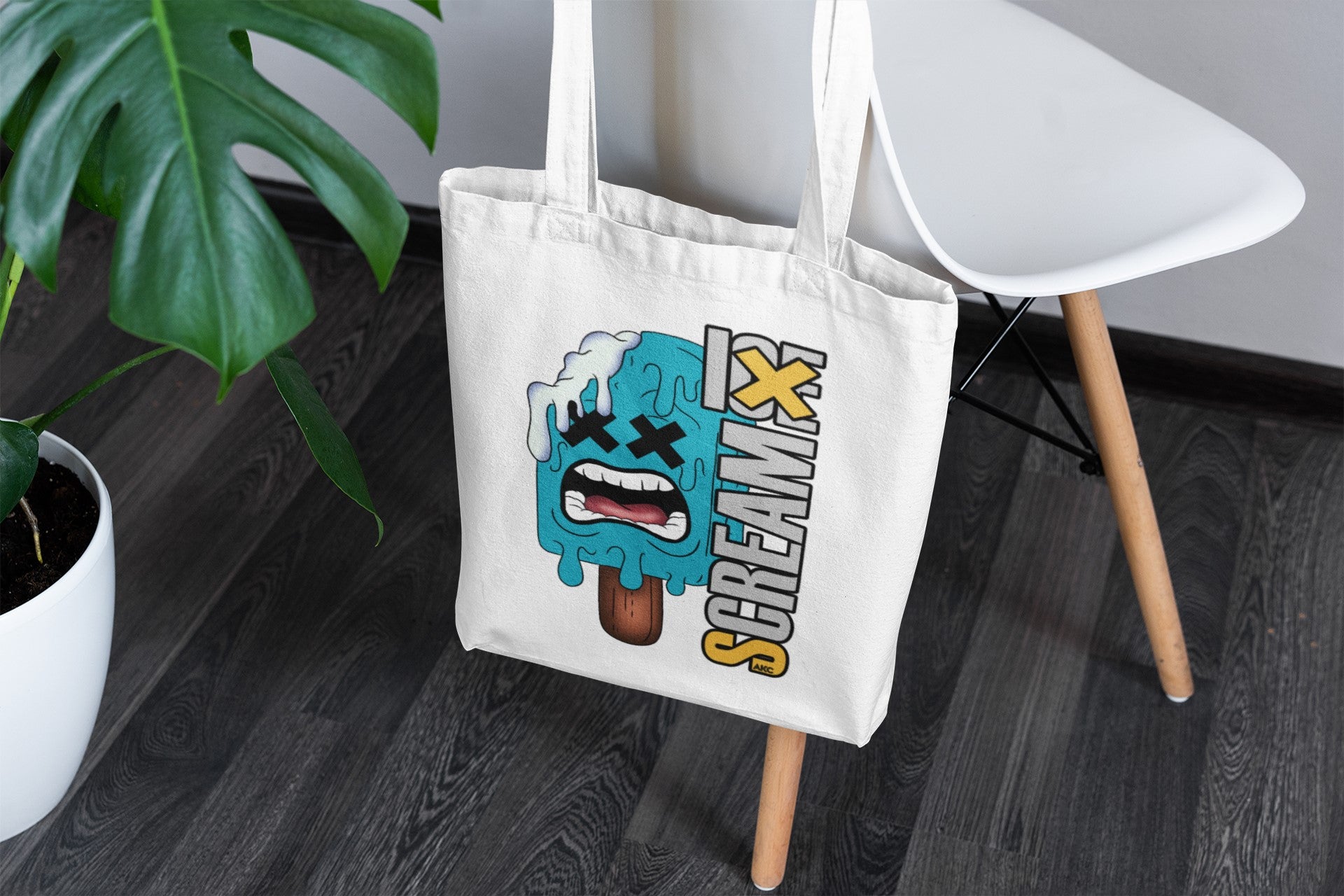 Tote bag with a graphic design on a wooden floor next to a plant and chair.