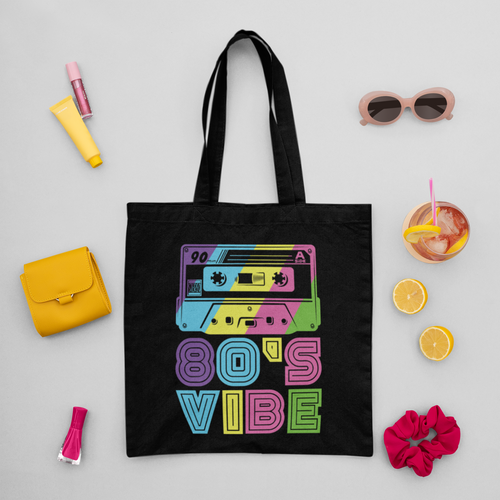 Black tote bag with '80s Vibe' design surrounded by small items on a light gray background