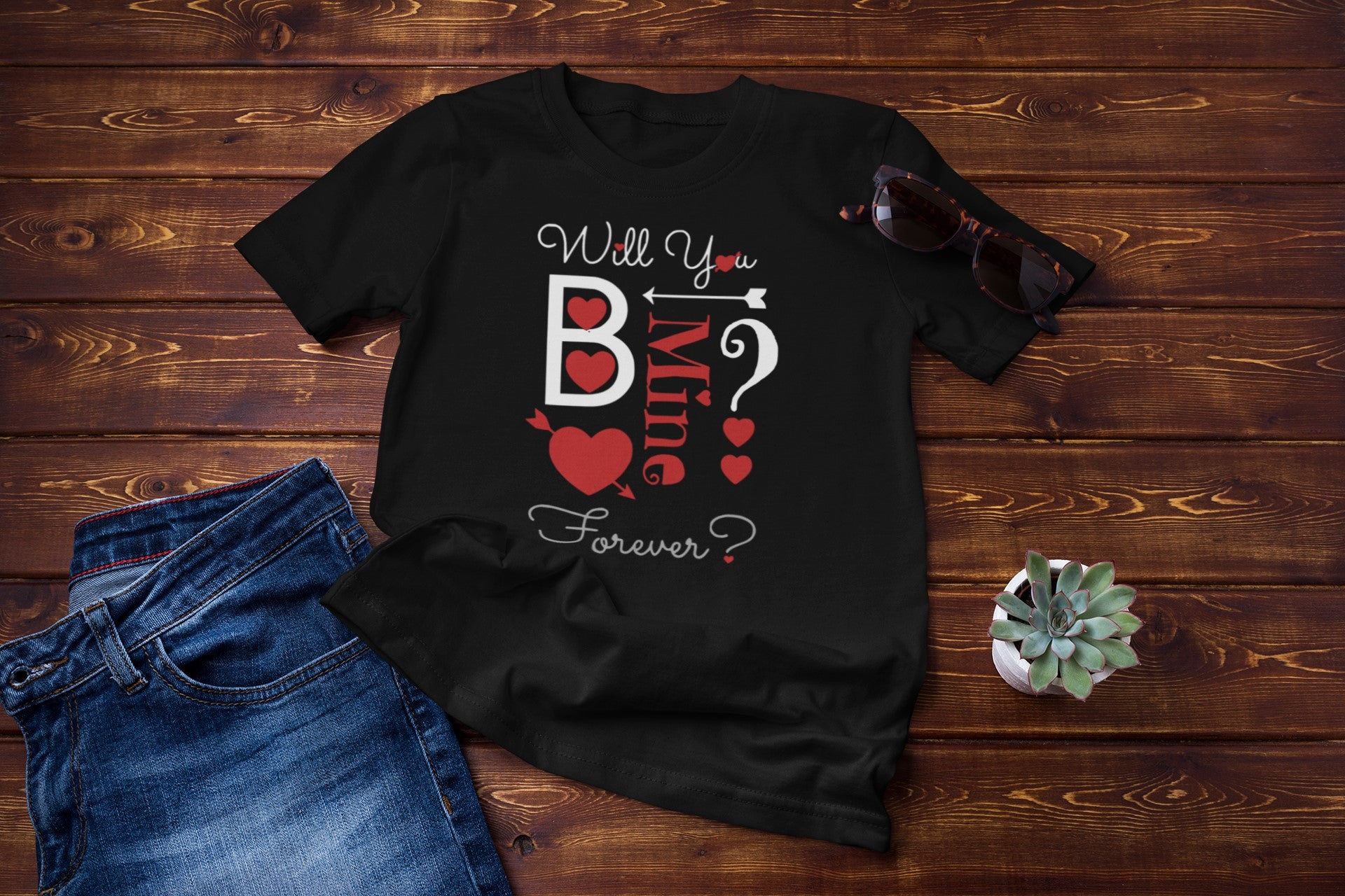Black t-shirt with 'Will You B My Valentine Forever?' text on a wooden surface with jeans and a plant.