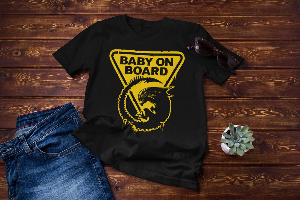 Black t-shirt with 'Baby on Board' graphic on a wooden surface with jeans and sunglasses.