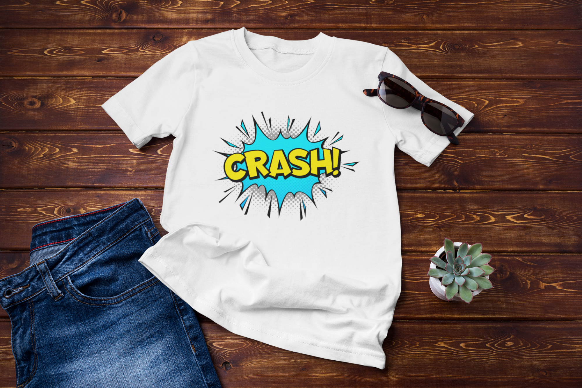 A white graphic t-shirt with a colorful comic-style 'CRASH!' speech bubble design, styled in a flat lay on a dark wood background with blue jeans, sunglasses, and a small succulent.