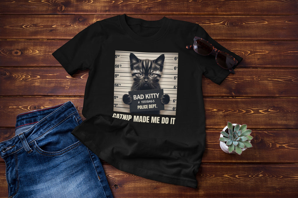 Black t-shirt with a graphic design of a cat and text, placed on a wooden surface with jeans and sunglasses.