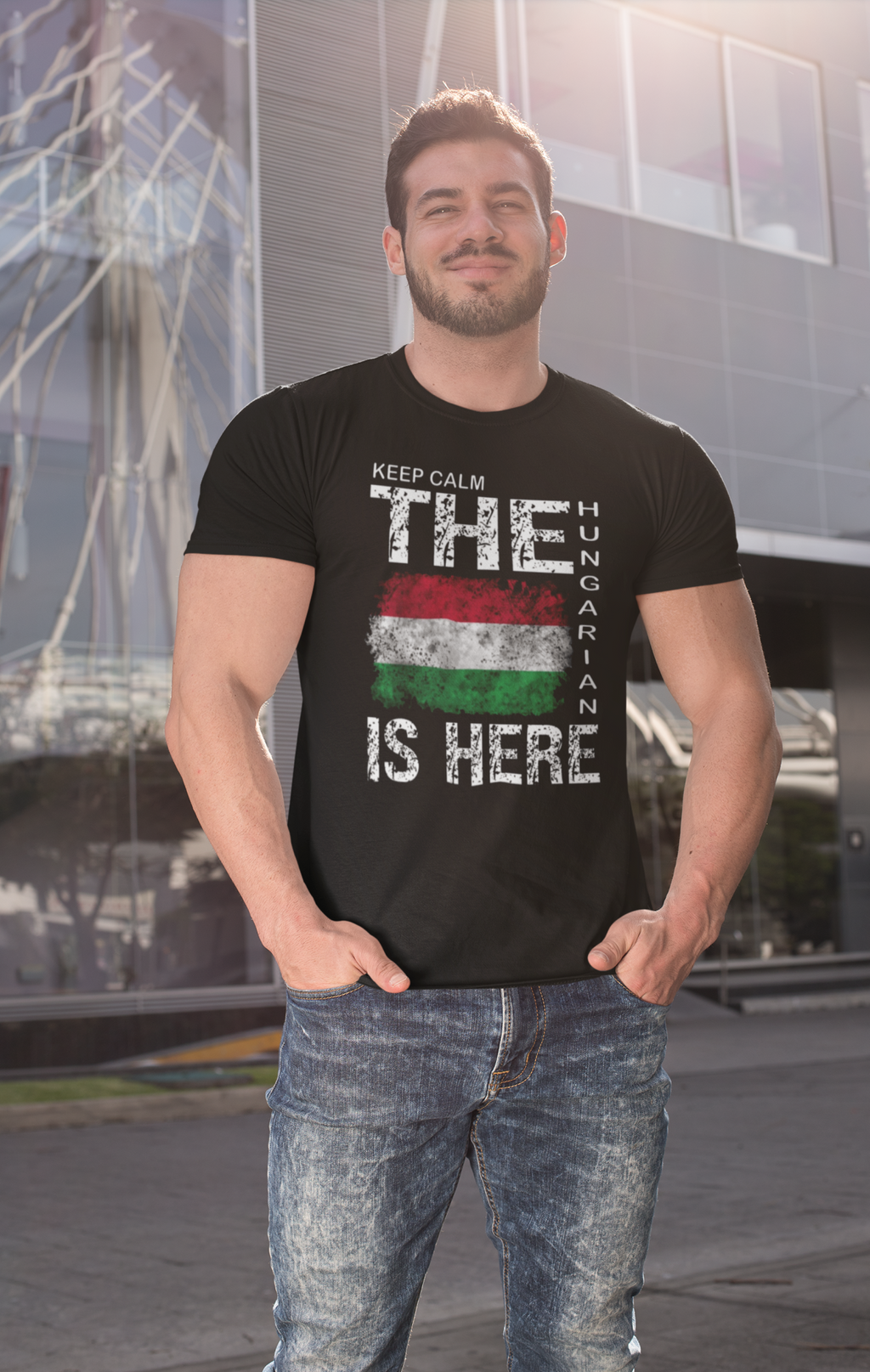 A man wearing a black graphic t-shirt with a distressed Hungarian flag and the text 'KEEP CALM THE HUNGARIAN IS HERE', paired with blue jeans, standing in front of a modern glass building.