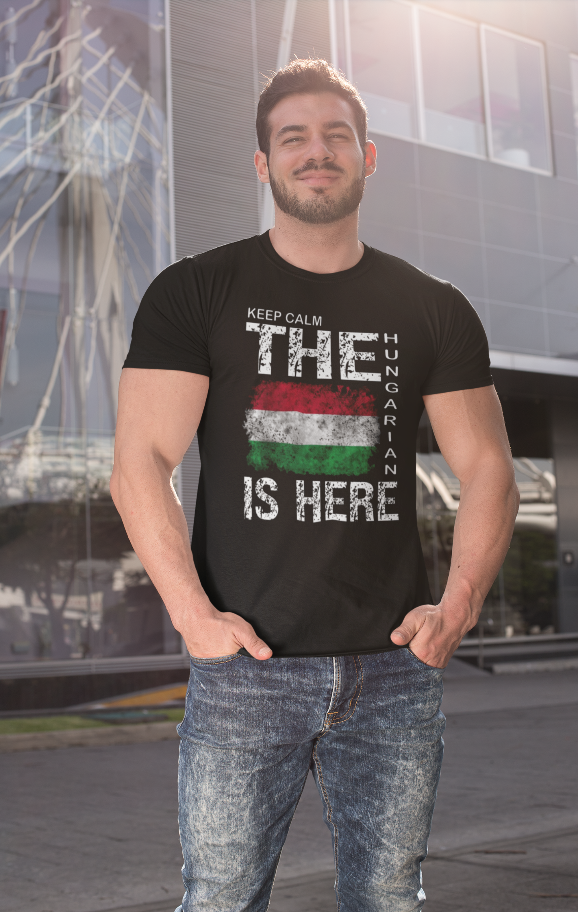 A man wearing a black graphic t-shirt with a distressed Hungarian flag and the text 'KEEP CALM THE HUNGARIAN IS HERE', paired with blue jeans, standing in front of a modern glass building.