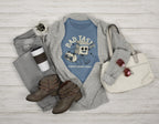 Blue t-shirt with graphic design, gray sweater, brown boots, white bag, and coffee cup on a wooden surface.
