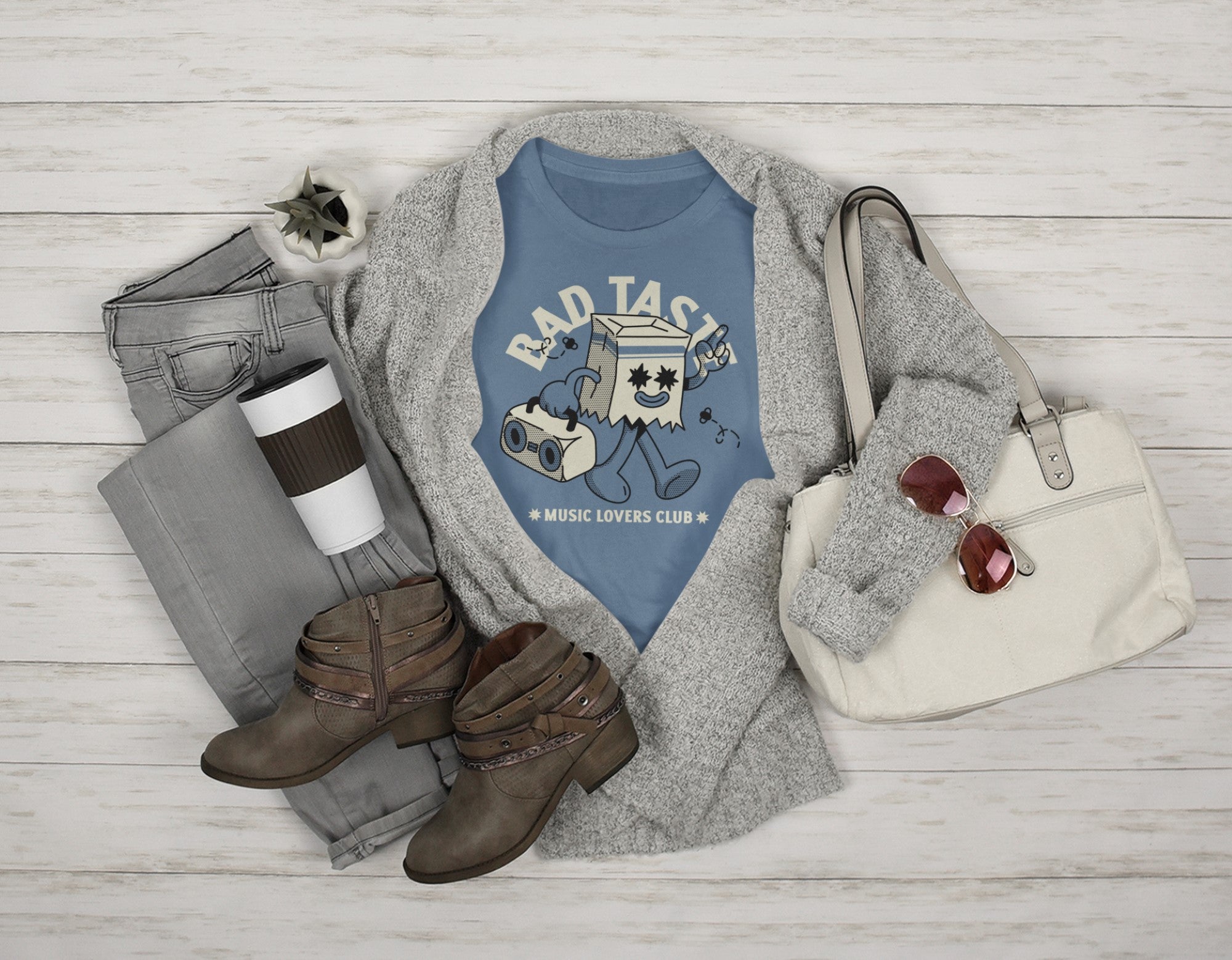Blue t-shirt with graphic design, gray sweater, brown boots, white bag, and coffee cup on a wooden surface.
