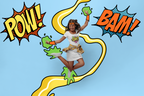A woman in a white t-shirt and leopard-print skirt jumps joyfully while wearing green slime and orange arm bands, set against a light blue background with comic-style 'POW!' and 'BAM!' speech bubbles.
