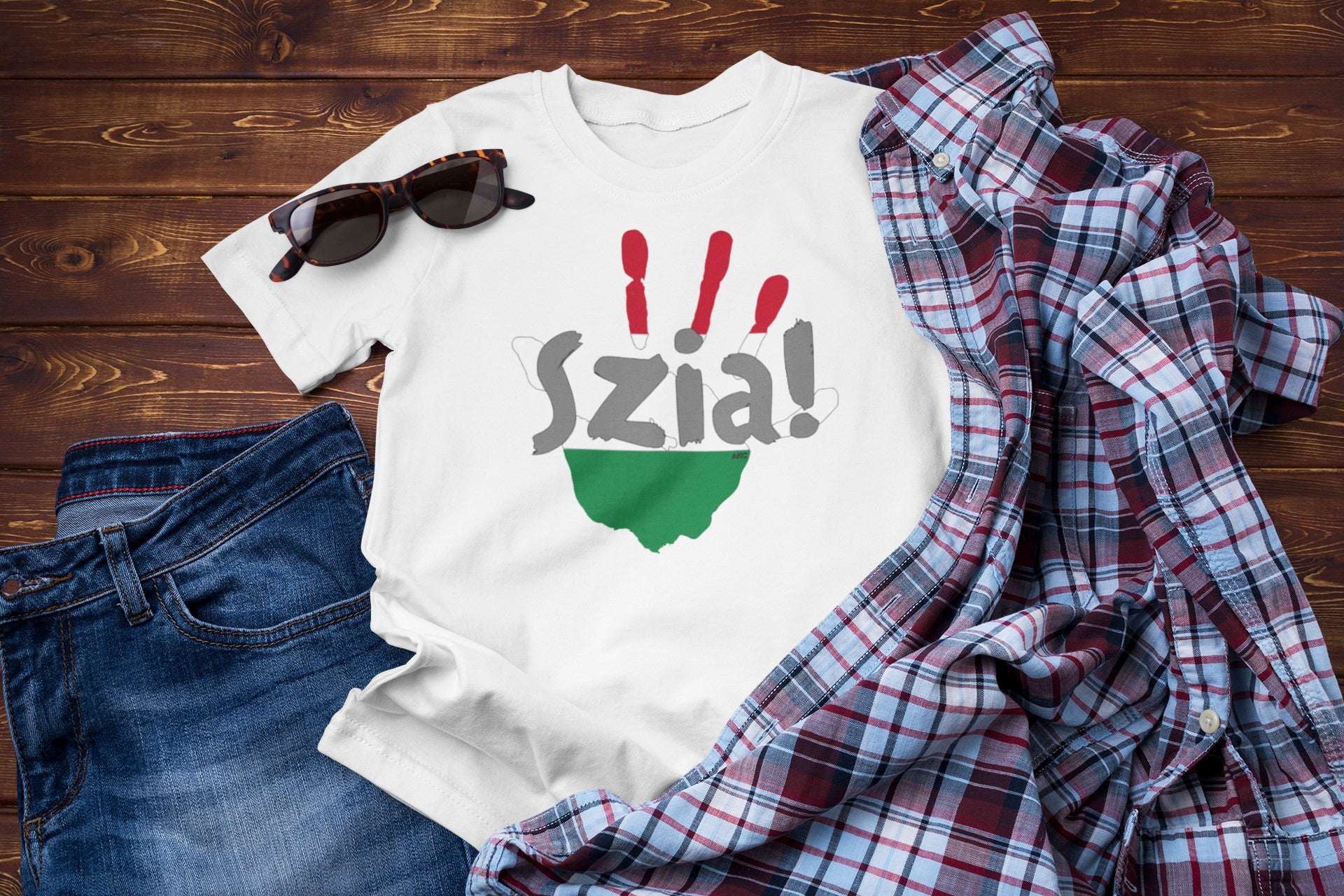 White t-shirt with 'Szia!' design on a wooden surface with jeans and a plaid shirt.