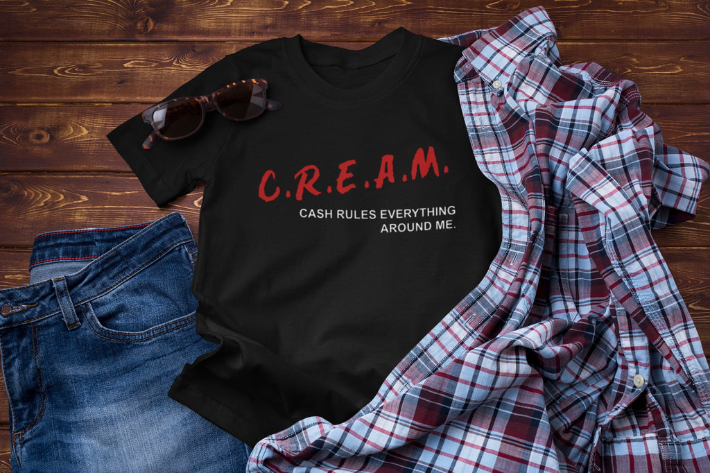 Black t-shirt with 'C.R.E.A.M. Cash Rules Everything Around Me' text on a wooden surface with jeans and a plaid shirt.