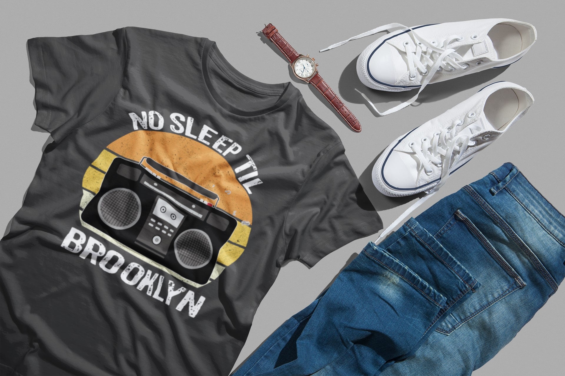 Gray t-shirt with 'No Sleep Till Brooklyn' design, white sneakers, blue jeans, and a watch on a gray background