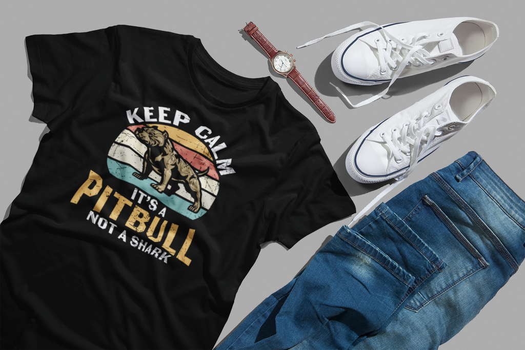 A black graphic T-shirt laid flat on a gray background, styled with white sneakers, blue jeans, and a brown watch. The shirt features a retro sunset graphic with a pitbull illustration and the text 'KEEP CALM IT'S A PITBULL NOT A SHARK'.