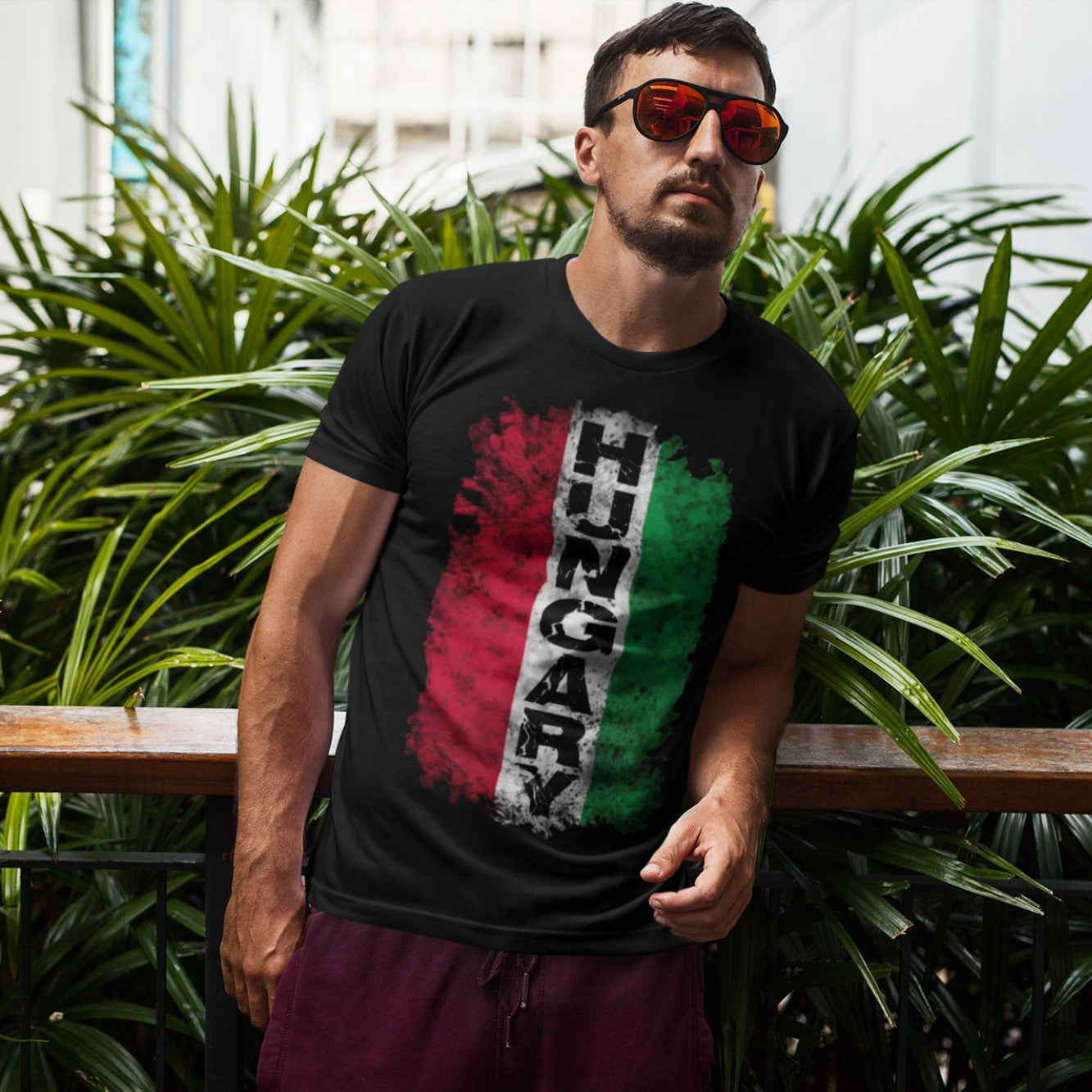 Man wearing a black t-shirt with Hungarian flag design and sunglasses, standing in front of green plants.
