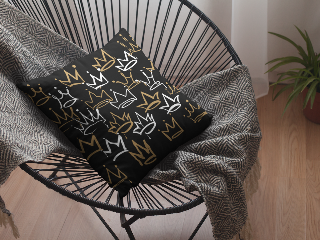 Decorative pillow with crown pattern on a black wire chair with a gray blanket.