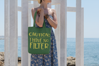 Person holding a green tote bag with 'CAUTION: I HAVE NO FILTER' text by a coastal background