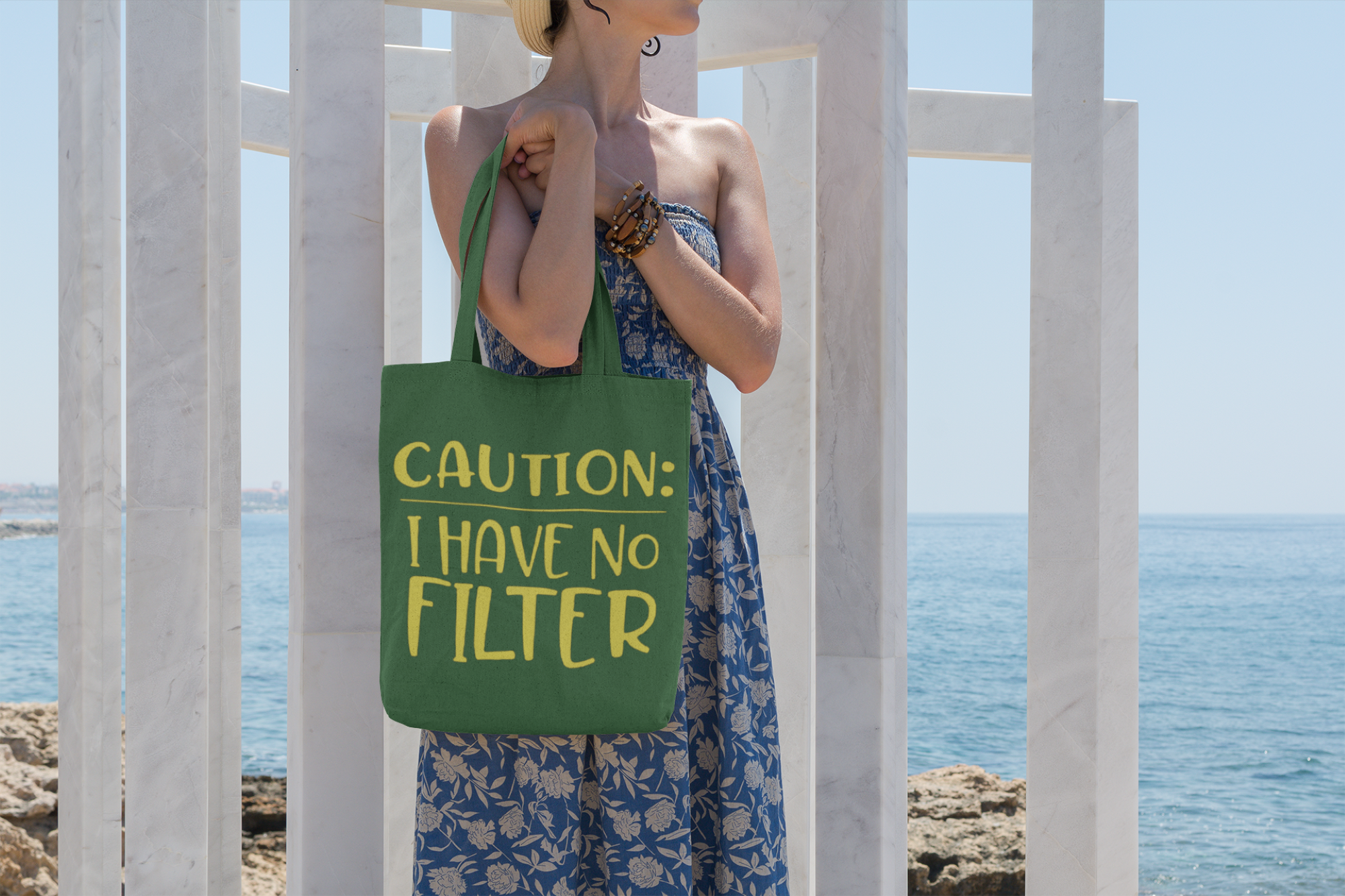 Person holding a green tote bag with 'CAUTION: I HAVE NO FILTER' text by a coastal background