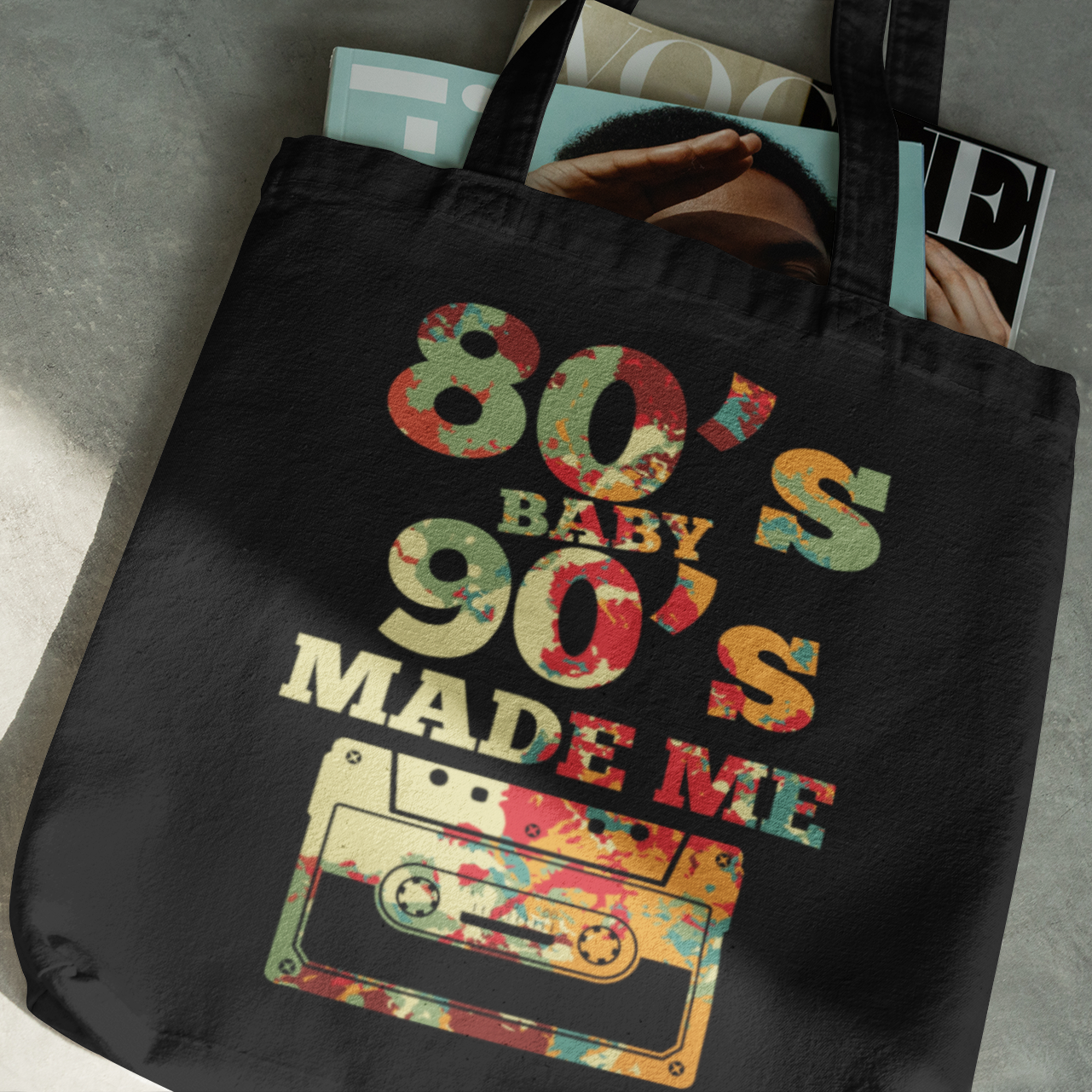 Black tote bag with '80s Baby 90s Made Me' text and cassette tape graphic on a gray surface.