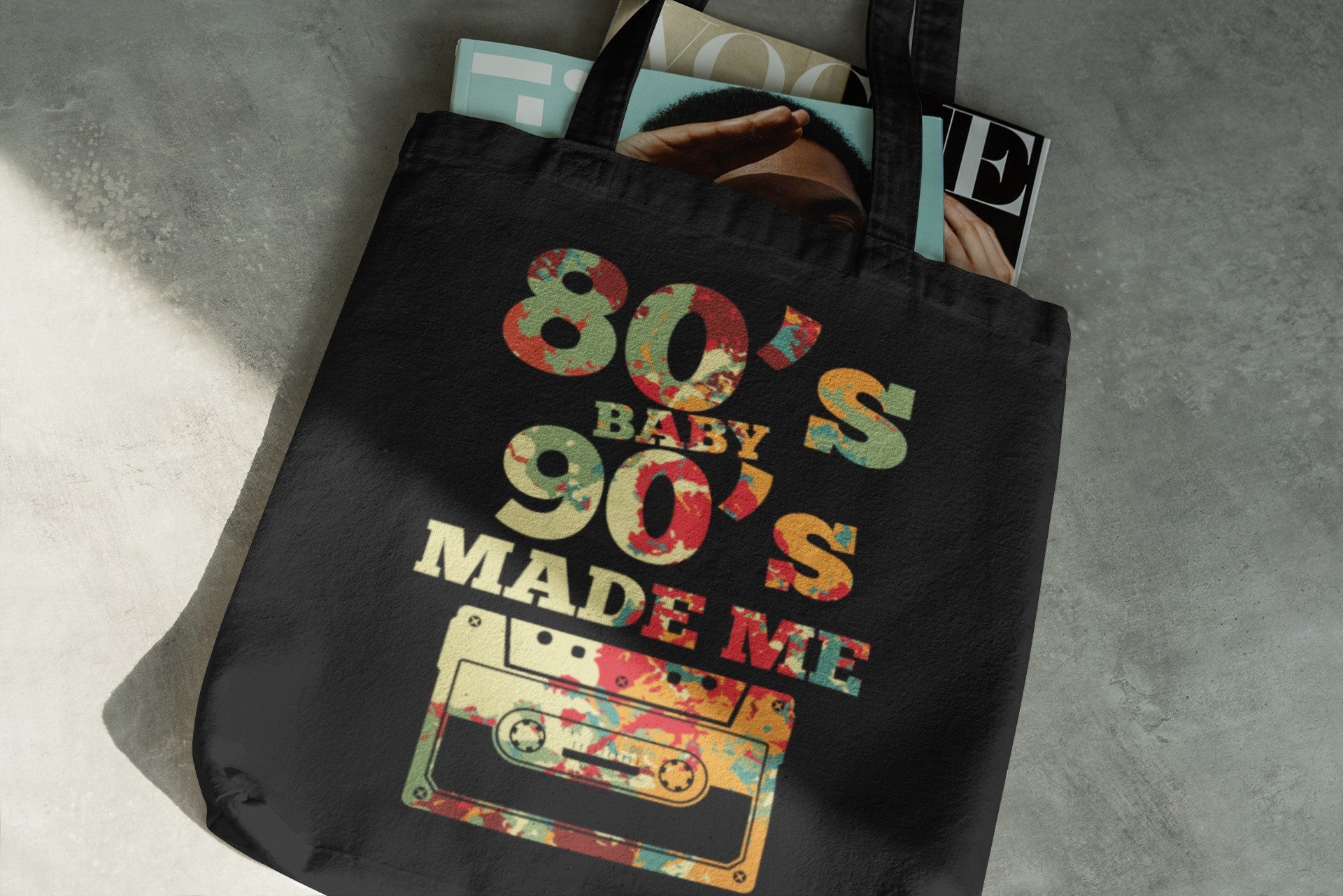 Black tote bag with '80s Baby 90s Made Me' design featuring a cassette tape graphic on a gray surface.