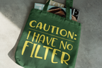 Green tote bag with yellow text 'CAUTION: I HAVE NO FILTER' on a gray surface.