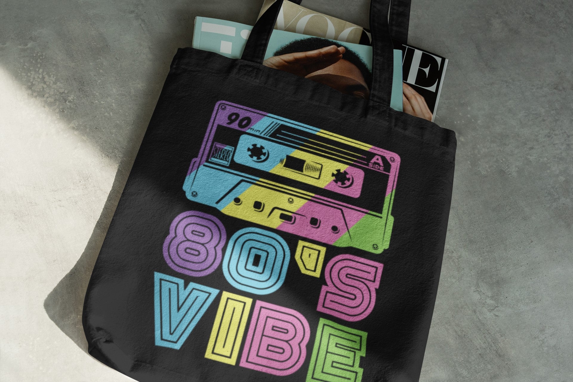 Black tote bag with colorful '80s Vibe' design featuring cassette tapes on a gray surface.
