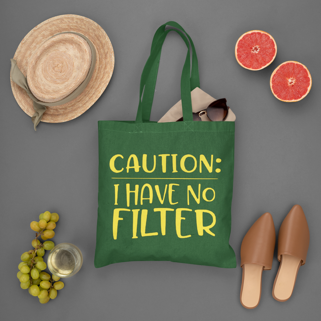 Green tote bag with 'CAUTION: I HAVE NO FILTER' text, surrounded by a hat, sunglasses, grapes, and shoes on a gray background.
