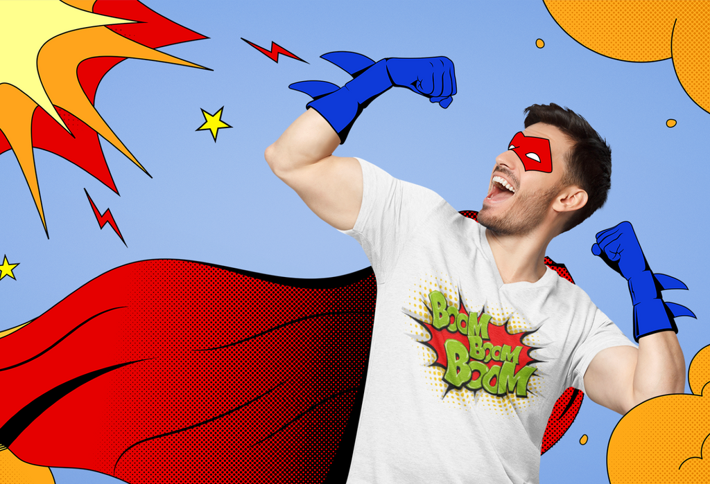 A man wearing a white graphic t-shirt with 'Boom Boom Boom!' printed on the front, styled as a comic book hero with a red cape, red mask, and blue gloves, set against a retro comic-style blue background with yellow clouds and stars.