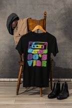 A black graphic t-shirt hanging on a wooden hanger, styled on a wooden chair with a tan jacket and dark gray hat draped behind it, with black ankle boots on the floor. The t-shirt features a colorful retro cassette tape graphic with the text '80'S VIBE' in neon pink, green, blue, and purple.