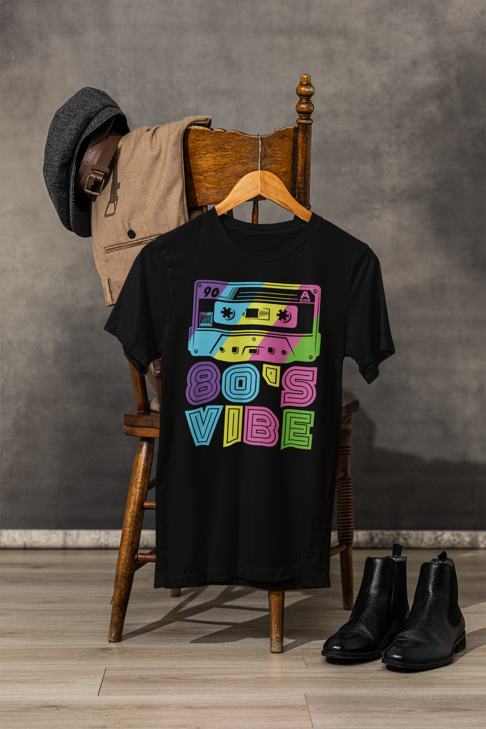 A black graphic t-shirt hanging on a wooden hanger, styled on a wooden chair with a tan jacket and dark gray hat draped behind it, with black ankle boots on the floor. The t-shirt features a colorful retro cassette tape graphic with the text '80'S VIBE' in neon pink, green, blue, and purple.
